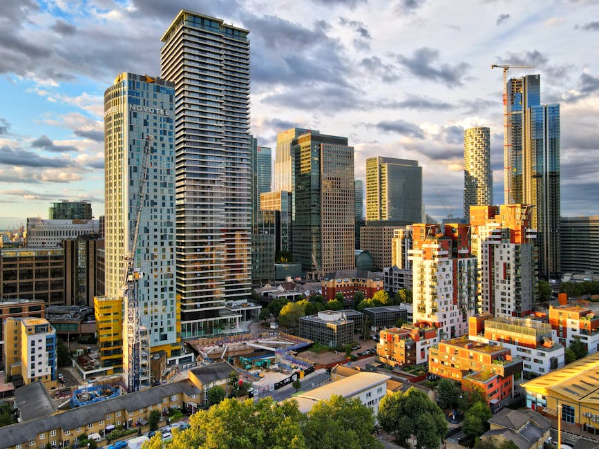 A wide-angle view of Canary Wharf in London during the early evening, featuring a cluster of modern high-rise buildings constructed with glass and steel, illuminated by interior and exterior lighting.