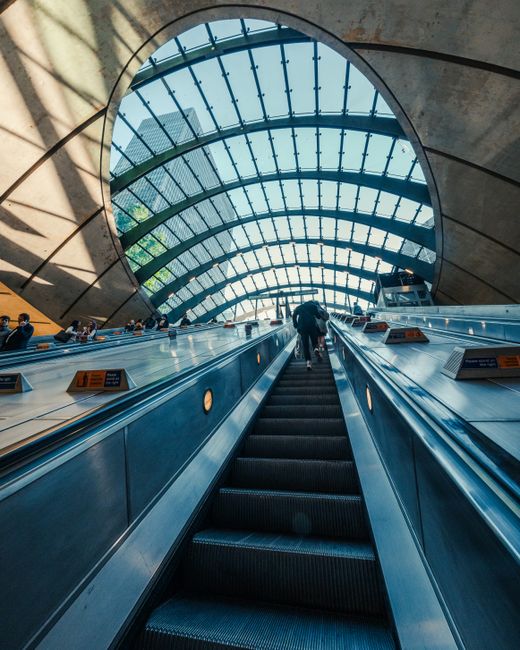 A modern indoor scene featuring a downward view along an escalator in a spacious building with a large, curved glass ceiling made of multiple panes supported by green metal beams. The escalator is fla