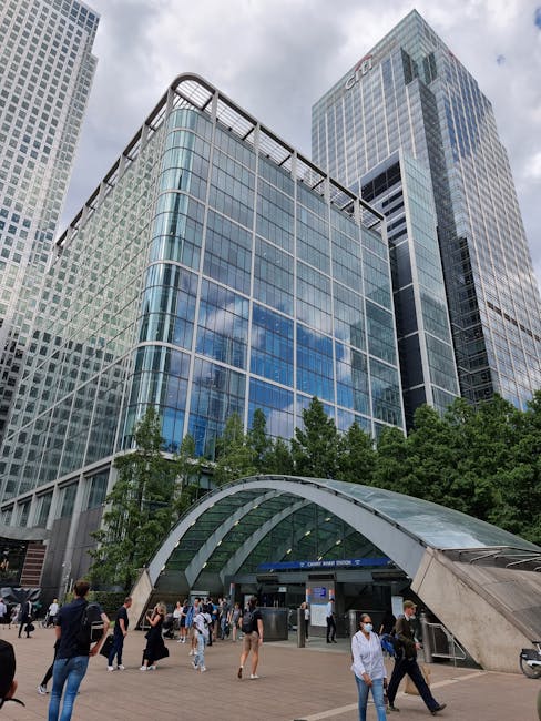 A low-angle view of modern office buildings in Canary Wharf, featuring glass and steel facades with reflective windows, set against a clear blue sky. The buildings showcase sleek, curved architectural