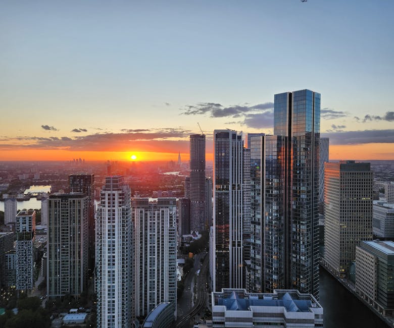 A panoramic view of a modern city skyline at sunset, featuring tall glass skyscrapers with reflective surfaces, some under construction with cranes, and surrounding high-rise buildings. The scene is i