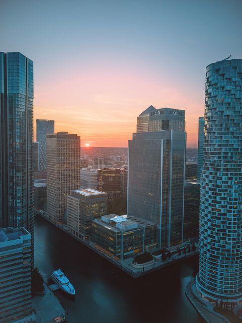 A collection of modern high-rise office buildings along a canal in Canary Wharf, featuring glass, steel, and concrete facades. The tallest building has a reflective glass exterior with a grid-like pat